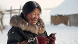 Smiling Eskimo woman wearing traditional clothing watching her mobile phone and laughing. Texting with smartphone in snow landscape