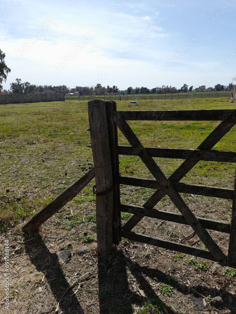 Puerta de madera o tranquera, cruzada en el campo de la Pampa Argentina, para la seguridad de los animales  con el sol de la mañana formando sombras de los postes, con un fondo verde y el cielo azul  