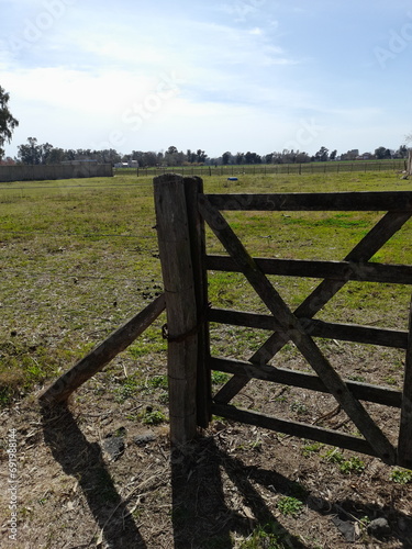 Puerta de madera o tranquera, cruzada en el campo de la Pampa Argentina, para la seguridad de los animales con el sol de la mañana formando sombras de los postes, con un fondo verde y el cielo azul 