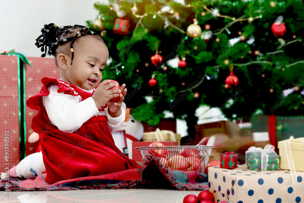 African little girl child eating apple and sitting on floor at living room with many present gift boxes under beautiful decorative Christmas tree. Cute kid celebrating happy Christmas winter holiday. African little girl child eating apple and sitting on floor at living room with many present gift boxes under beautiful decorative Christmas tree. Cute kid celebrating happy Christmas winter holiday.