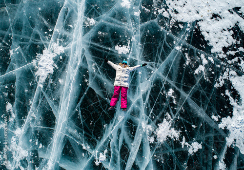 Aerial top down shot of the young woman lying on the blue cracked ice of Baikal and shaking hands to the camera. Popular tourist spot. Winter landscape of frozen Baikal photo