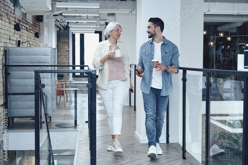 Smiling man ans woman holding coffee while walking in the office hall, talking, having coffee break. Full-length. Teamwork, startup, business concept