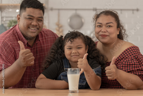 girl is holding a glass of milk. They were happily inviting their girl to drink morning milk together in the kitchen of their home. Breakfast time of Asian dad mom and kid people.
