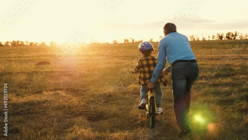 Affectionate papa actively involved in assisting little kid on riding bicycle. Caring dad prevents mishaps holding bike of son. Father provides risk-free bike ride for beloved child in field photo