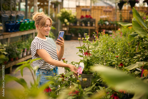 Woman Inside Greenhouse In Garden Centre Taking Picture Of Red Echinacea Plant On Mobile Phone