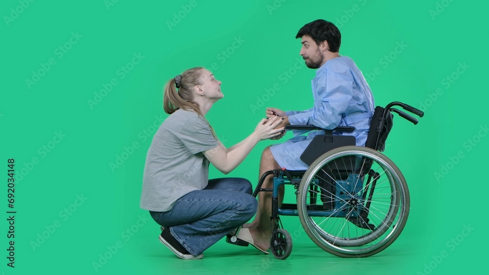 Adult man patient in robe sitting in wheelchair girl next to him on knees holding his hand, supporting expression. Isolated on chroma key green screen. Adult man patient in robe sitting in wheelchair girl next to him on knees holding his hand, supporting expression. Isolated on chroma key green screen.