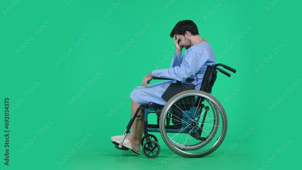 Obraz premium Adult man patient in robe sitting in wheelchair holding head in hand, looking at his legs, depressed expression sad face. Isolated on chroma key green screen.