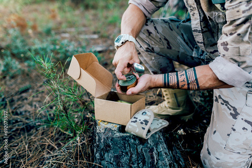 Anonymous Military soldier with backpack opening food can kneeling by box on tree photo