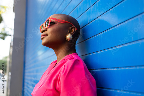 Happy Stylish black woman with shaved head in sunglasses and pink dress standing against blue wall photo