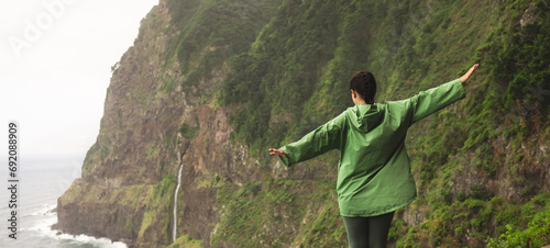 Back view of carefree unrecognizable young woman in green raincoat standing with arms outstretched on concrete barrier against mountains