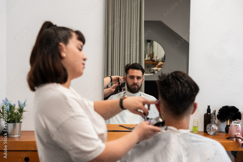 Side view of focused female hairstylist using electric trimmer on male client's hair at salon with mirror Side view of focused female hairstylist using electric trimmer on male client's hair at salon with mirror