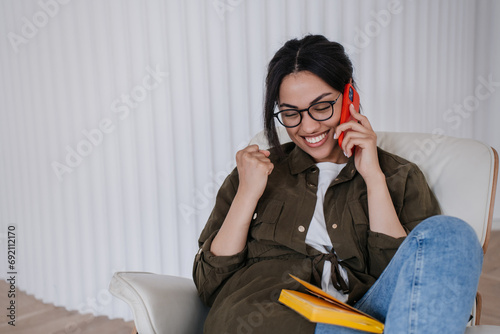 Adorable African american girl in glasses holding diary, speaking by phone, clenches fist, toothy smiling, celebrates success. Great deal. Entrepreneur satisfied by month profit. Success concept. photo