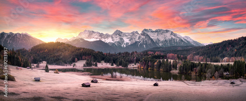 Stunning sunrise scenery with mountains, a scenic lake and woods on hills in the Alps, frozen meadow and cabins, beautiful nature panoramic shot photo
