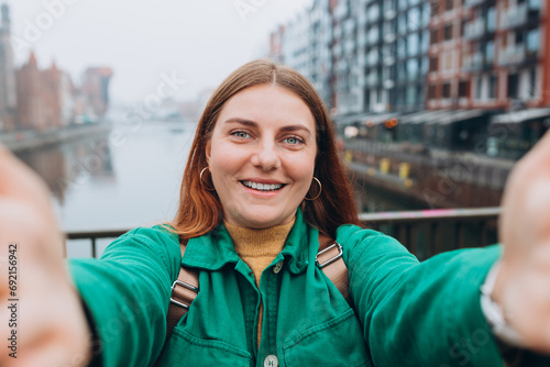 Happy 30s Women taking selfie on urban background. Young beautiful girl say Hi. Gdansk old town and famous Zuraw crane, A beautiful redhead woman is standing near Motlawa river, Traveler photo