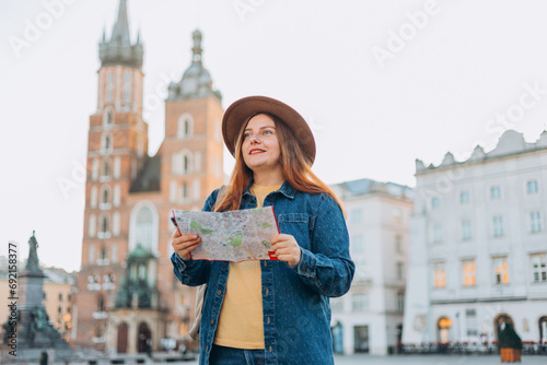 Attractive young female tourist is exploring new city. Redhead girl holding a paper map on Market Square in Krakow. Traveling Europe in autumn. St. Marys Basilica. Vacation concept © mdyn