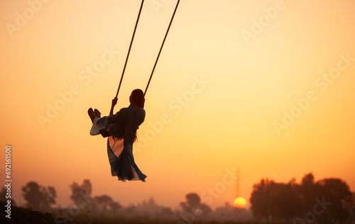 silhouette of a child playing on a swing. A young girl swinging at village during sunset
