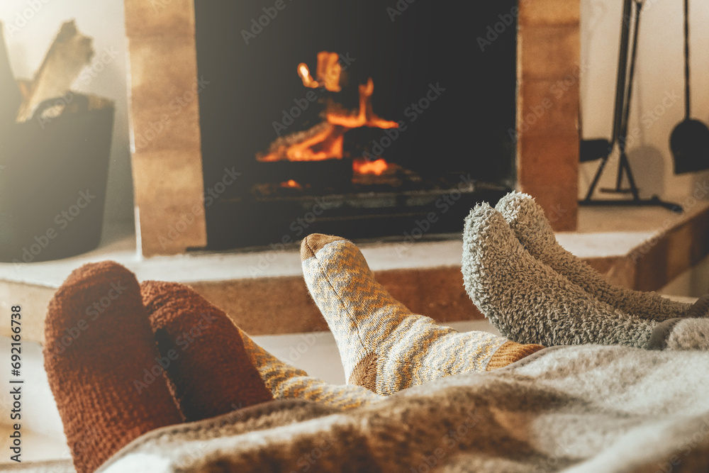 Family resting feet by fireplace on fall or winter time. People warming feet with blanket and socks by cozy fire. - obrazy, fototapety, plakaty 