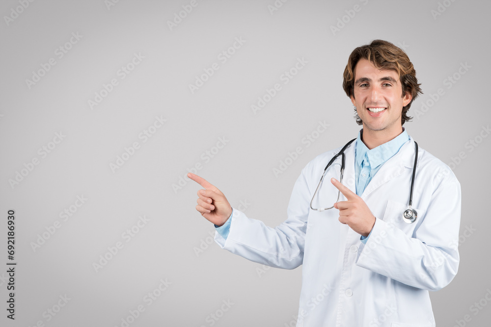 Happy young male doctor point fingers to empty space, isolated on grey background, smiling at camera Happy young male doctor point fingers to empty space, isolated on grey background, smiling at camera