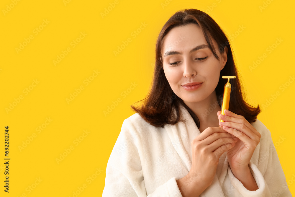 Young woman in bathrobe with facial massage tool on yellow background, closeup Young woman in bathrobe with facial massage tool on yellow background, closeup