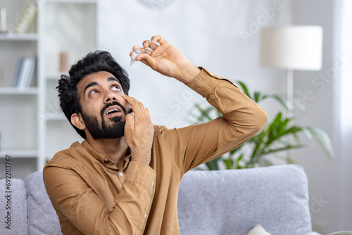 A young Indian man sits at home sick on the sofa and drops medicine in his eyes.