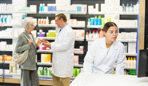 Girl pharmacist in pharmacy works with computer, searches among nomenclature of drugs, specifies price and availability of medicine photo