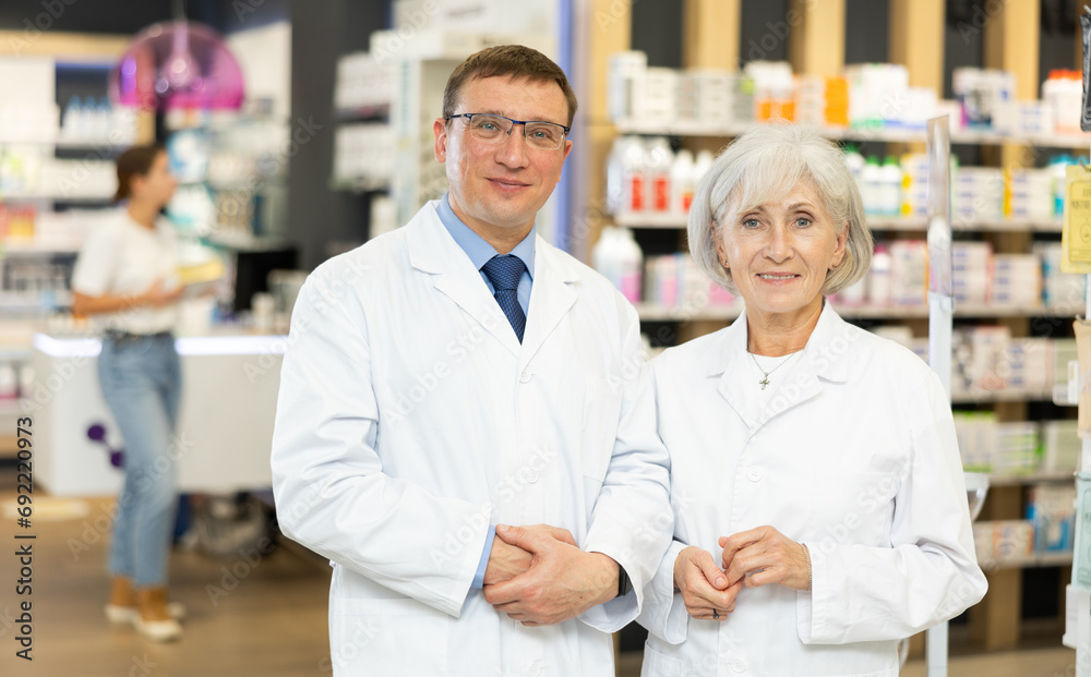 Senior woman and man pharmacists are standing in pharmacys sales hall, pharmacy staff are amicably preparing to meet chemist shop visitors