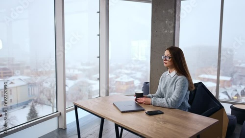 Thoughtful Caucasian woman wearing glasses sitting at desk in office. Lady takes her phone in hands, looks at screen and attaches gadget to her ear. photo