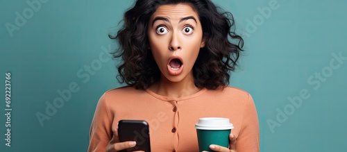Beautiful hispanic woman using smartphone and drinking a cup of coffee in shock face looking skeptical and sarcastic surprised with open mouth. Copy space image. Place for adding text or design photo