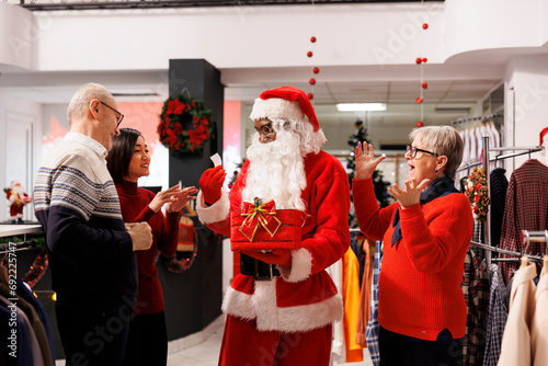 Young man presenting contest winners in clothing store, reading names of participants from papers. African american person in santa claus costume greeting people at prize drawing raffle. photo