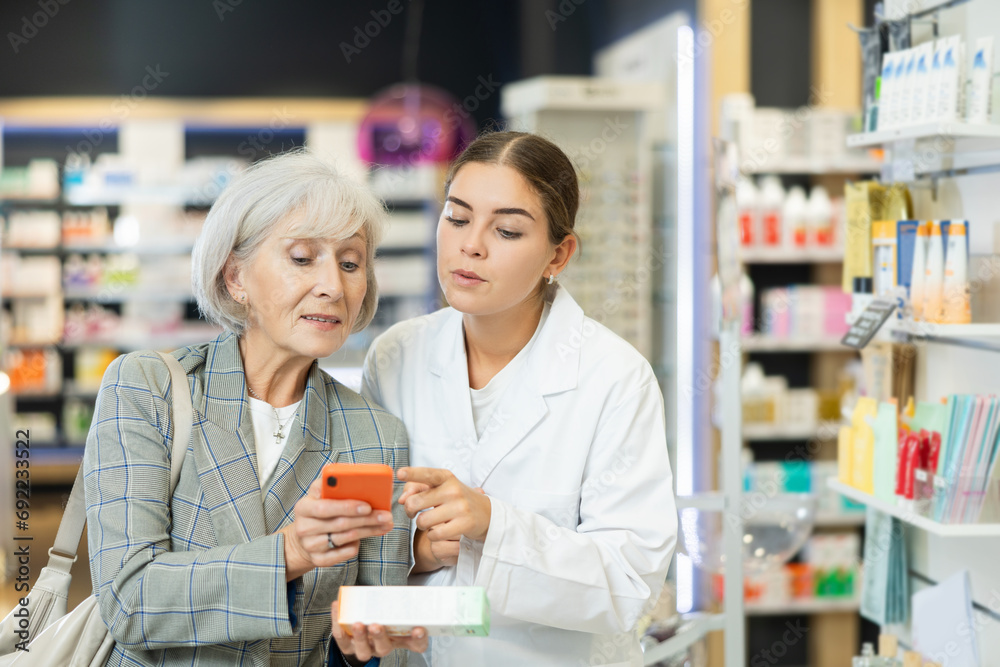 Concerned old woman showing to young female pharmacist QR-code of product on her smartphone in chemist's shop