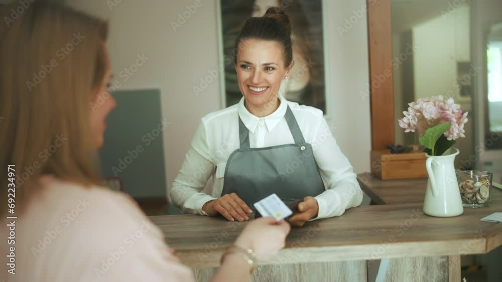 40 years old hair salon employee in modern beauty studio with client accepting credit card payments via terminal on reception.