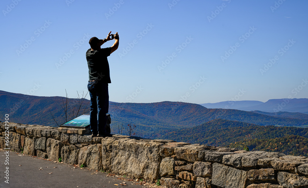 Obraz premium A visitor to Skyline Drive in VA stands on the stone wall to get a better shot of the changing Autumn Leaves and the distant ridge with his cell phone.