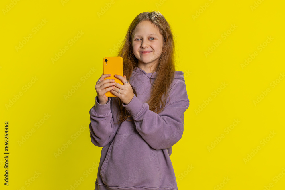Smiling preteen child girl kid using mobile smartphone typing new post on web, looking at camera, sms message, watching video addiction of social media networks. Teenager children on yellow background Smiling preteen child girl kid using mobile smartphone typing new post on web, looking at camera, sms message, watching video addiction of social media networks. Teenager children on yellow background