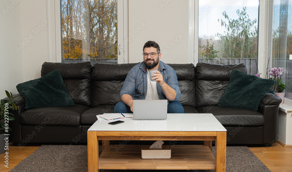 Happy man drinking coffee using a laptop sitting on a comfortable couch at home in the living room. Happy man drinking coffee using a laptop sitting on a comfortable couch at home in the living room.