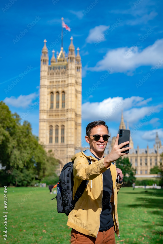 Obraz premium Cheerful young Latin man in casual clothes with backpack in sunglasses and jacket smiling and taking selfie while standing on background of Big Ben against clock tower and cloudy sky in London