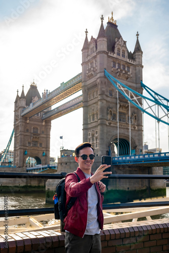 Happy young Latin man wearing sunglasses and red jacket with backpack taking selfie with smartphone near metal railing of famous Tower Bridge in London