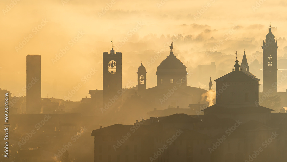 Fototapeta premium Bergamo, Italy. Amazing aerial landscape of the fog rises from the plains and covers the old town during sunrise. Warm colors. Bergamo one of the most beautiful city in Italy