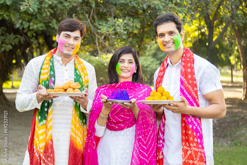 Happy group of three indian people wearing white kurta holding colorful paint or gulal plate in their hands celebrating holi festival at park or garden.
