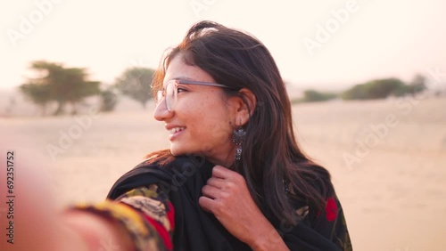 Happy teen girl taking selfie video on desert background while standing at Khuri sand dunes in Rajasthan, India. Indian woman social media influencer  streaming. POV of Indian woman in outdoors. photo