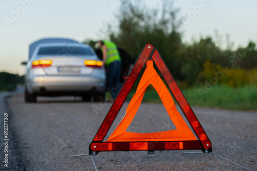 Unrecognizable sad driver in reflective vest. Male driver standing near broken car with open up hood. Red triangle to warn other road users of car breakdown or engine failure stop at countryside