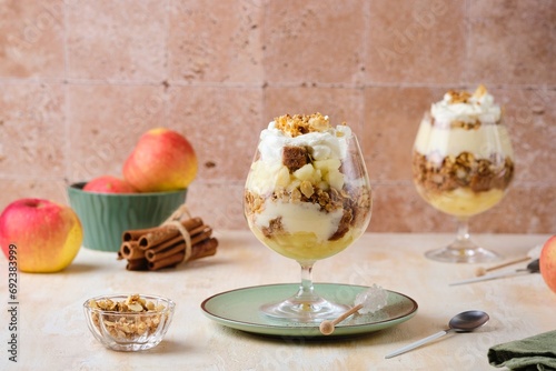 Dessert, portioned apple trifle with oat crumble, spiced cake, custard and whipped cream in a glass goblet on a light concrete background. photo