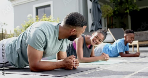 Happy african american father, son and daughter exercising in garden doing plank, slow motion photo