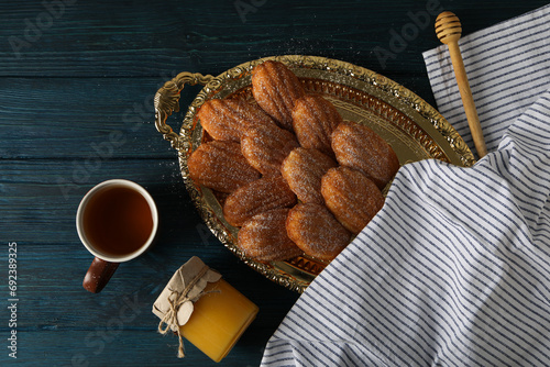 Madeleine cakes, cup of tea, jar of honey and towel on blue wooden background, top view photo
