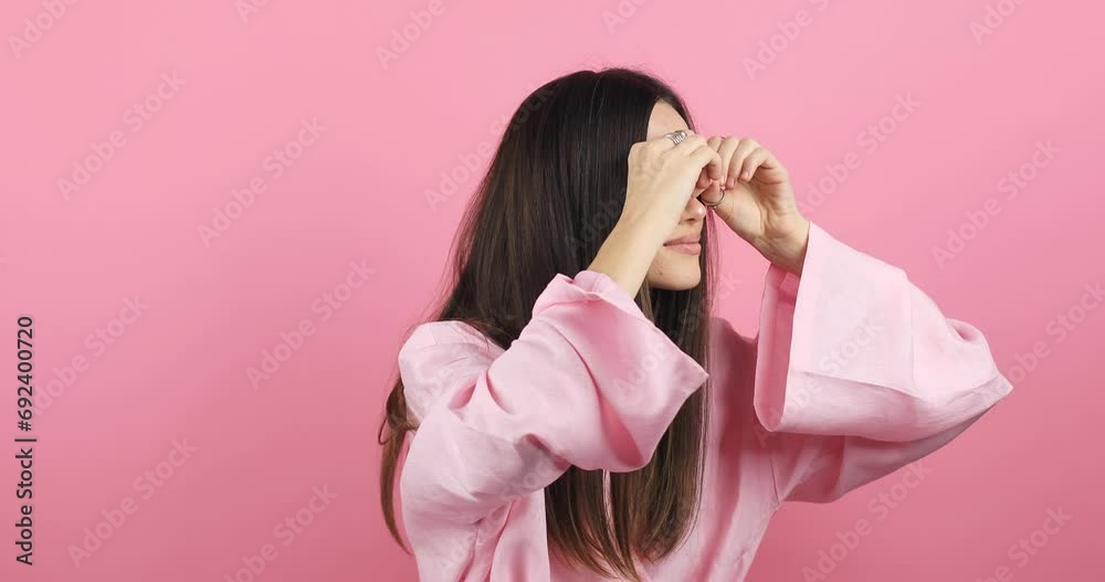 Amazed curious woman with brunette hair in pink dress looking around through fingers imitating binoculars and point at something surprised. Indoor studio shot isolated on pink background. Amazed curious woman with brunette hair in pink dress looking around through fingers imitating binoculars and point at something surprised. Indoor studio shot isolated on pink background.