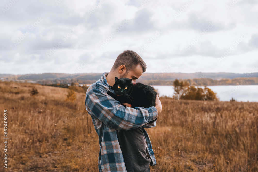 A Loving Man and His Black Cat in the Autumn Forest. He Carries His Pet on His Shoulder and Walks with a Leash in the Park. A Loving Man and His Black Cat in the Autumn Forest. He Carries His Pet on His Shoulder and Walks with a Leash in the Park.