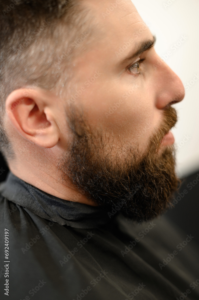 Obraz premium A young man with a beard in a black cape sits on a chair in a barbershop. The client is waiting for a haircut of the beard.