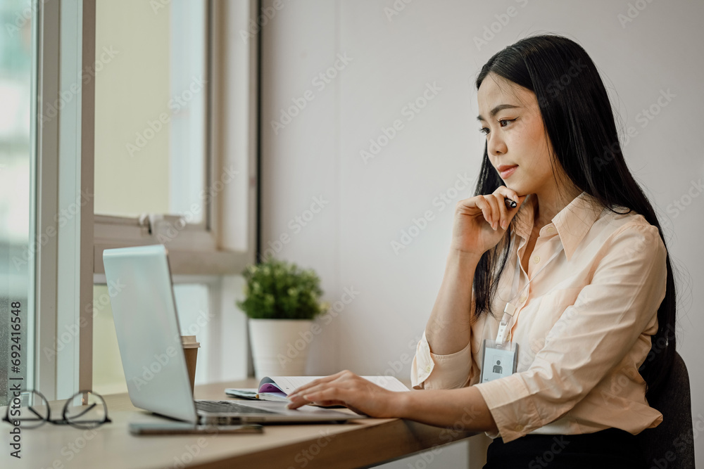 Focused Asian female employee looking at laptop screen, reading email at desk.