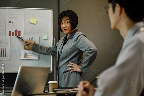 Serious senior businesswoman standing in front of flip chart explaining task at meeting.