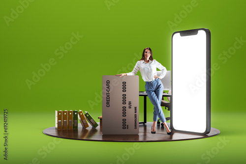 Smiling young woman standing with big credit card near giant 3D model of mobile phone with empty screen over green background. Online shopping photo