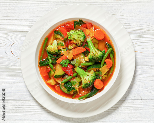 vegetable soup with green beans, broccoli, carrot, celery, spinach, cabbage, zucchini, tomatoes in white bowl on white wooden table, horizontal view, close-up, flat lay